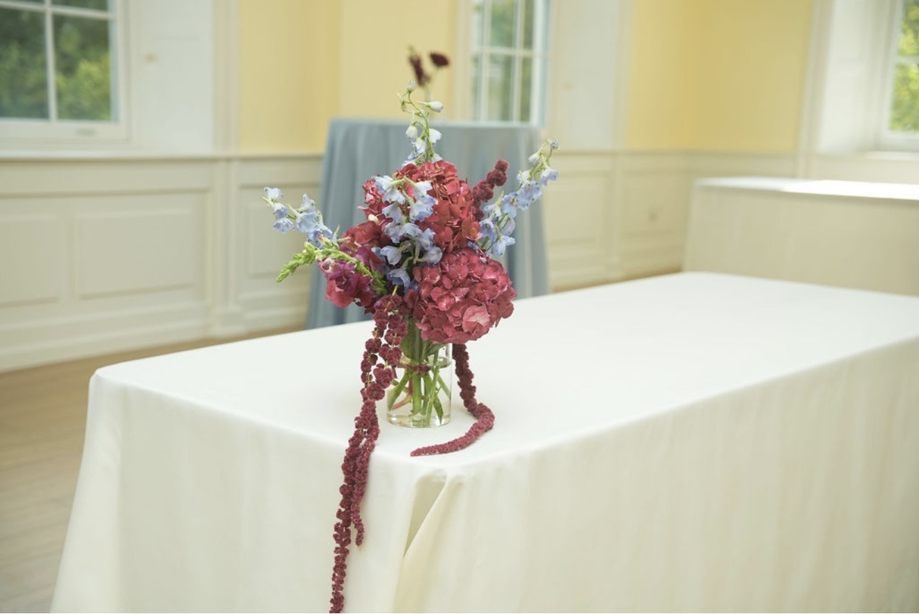 A single arrangement of burgundy hydrangea, pale blue delphinium and trailing amaranthus on an otherwise bare ivory table.