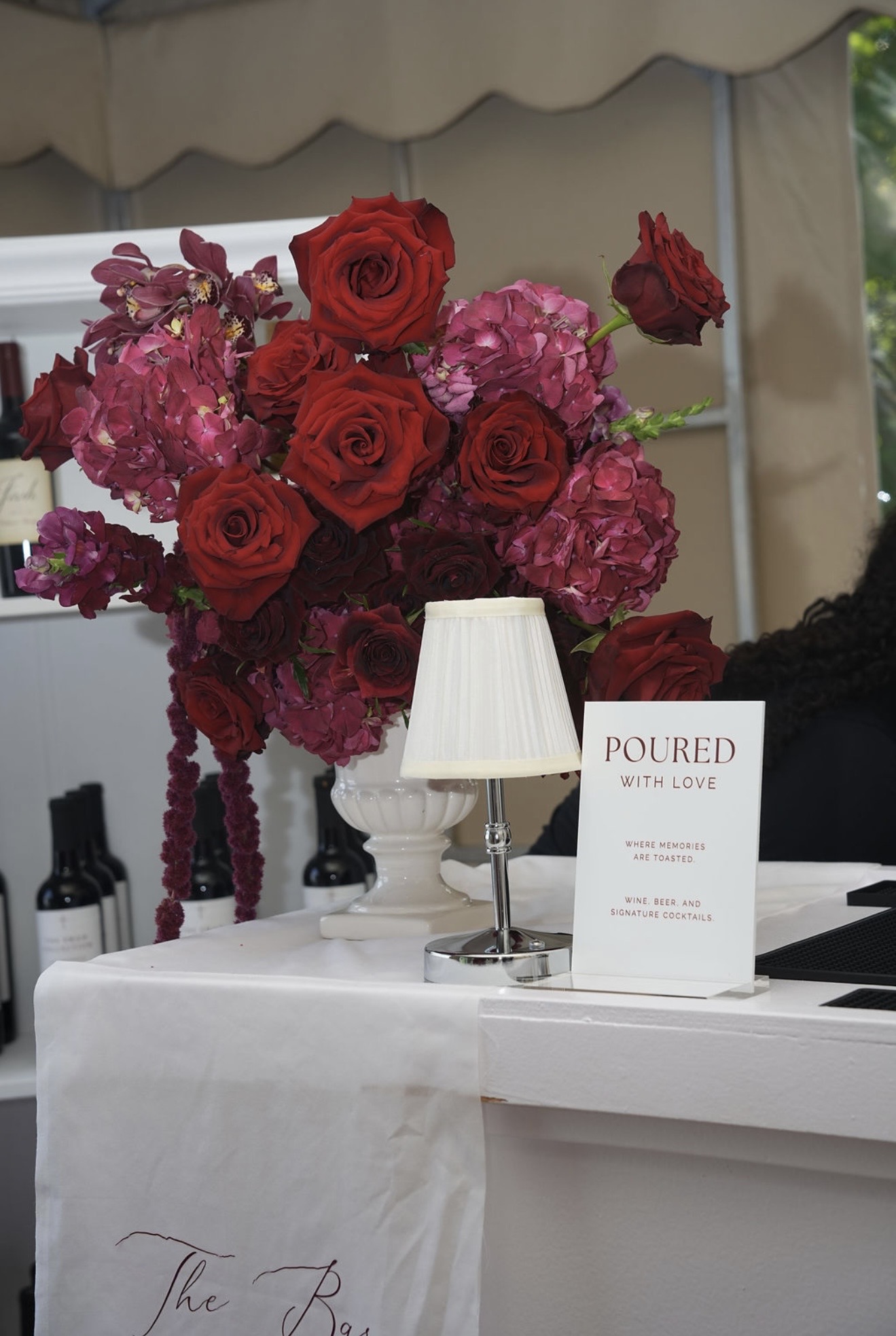 A tall arrangement of red roses, orchids and hydrangea on the bar, beside a shaded lamp and a card reading Poured With Love.