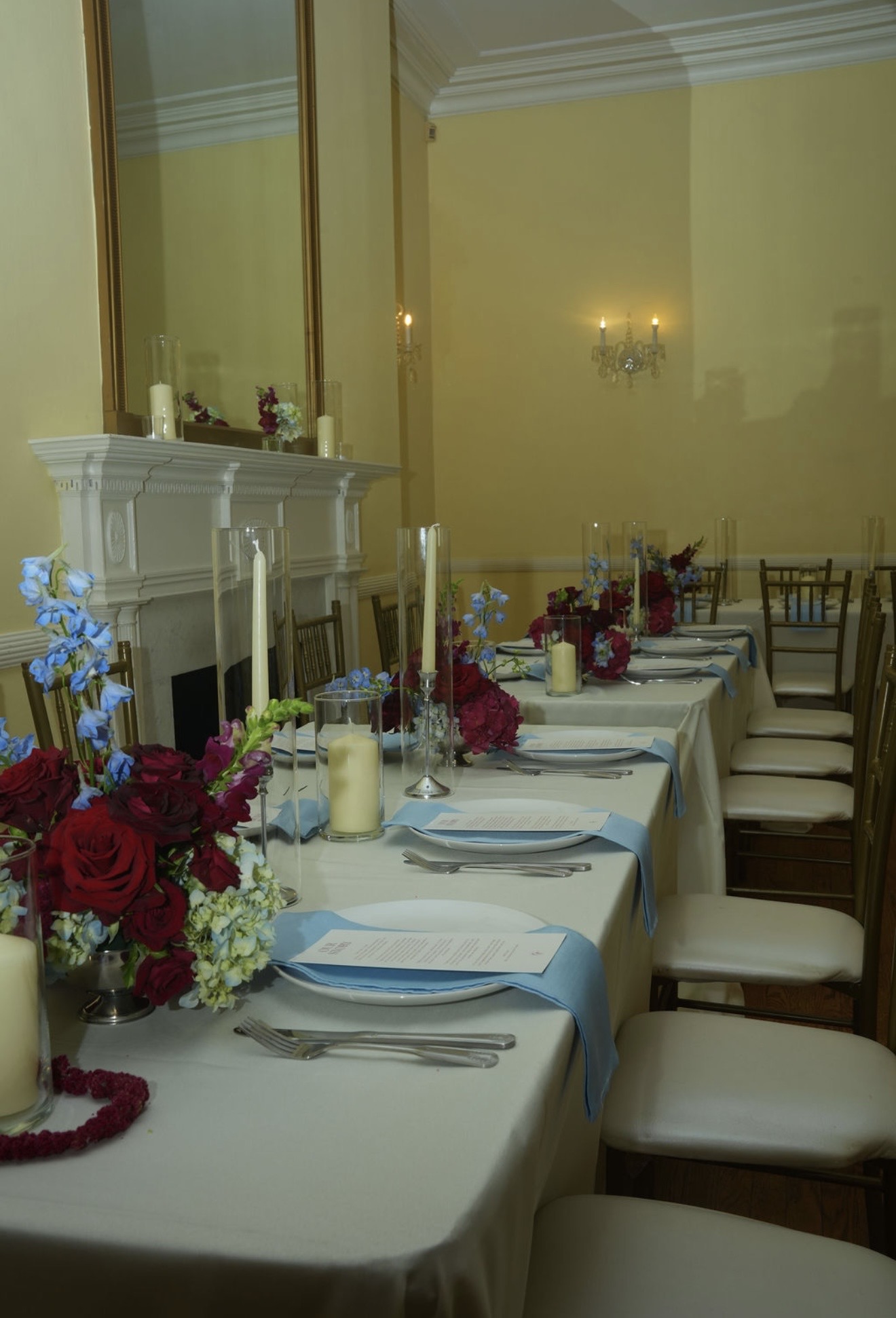 A long ivory table set with taper candles, pillar candles in glass, red and blue florals, pale blue napkins and printed menus, running toward a white mantel.