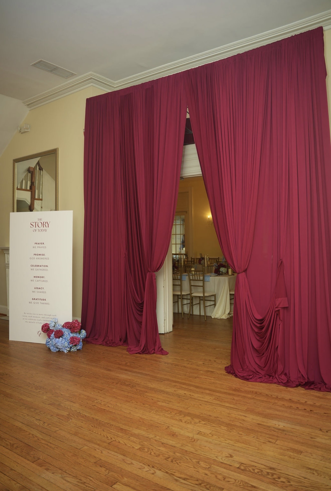 Floor-to-ceiling crimson drapery parted at a doorway, with a printed sign titled The Story of Today and a low arrangement of red and blue hydrangea beside it.