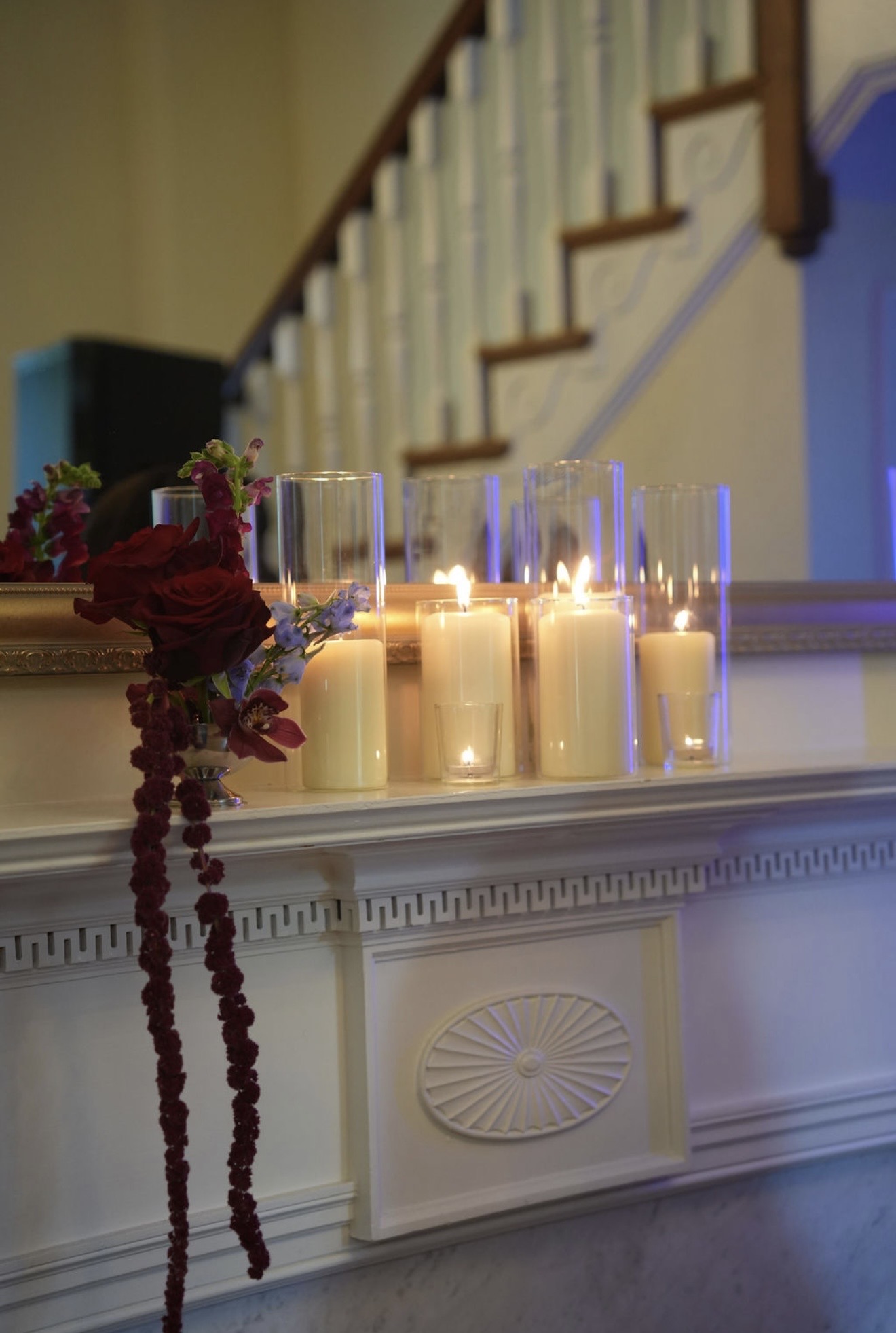 Pillar candles in glass hurricanes along a white mantel, with deep red roses and trailing amaranthus.