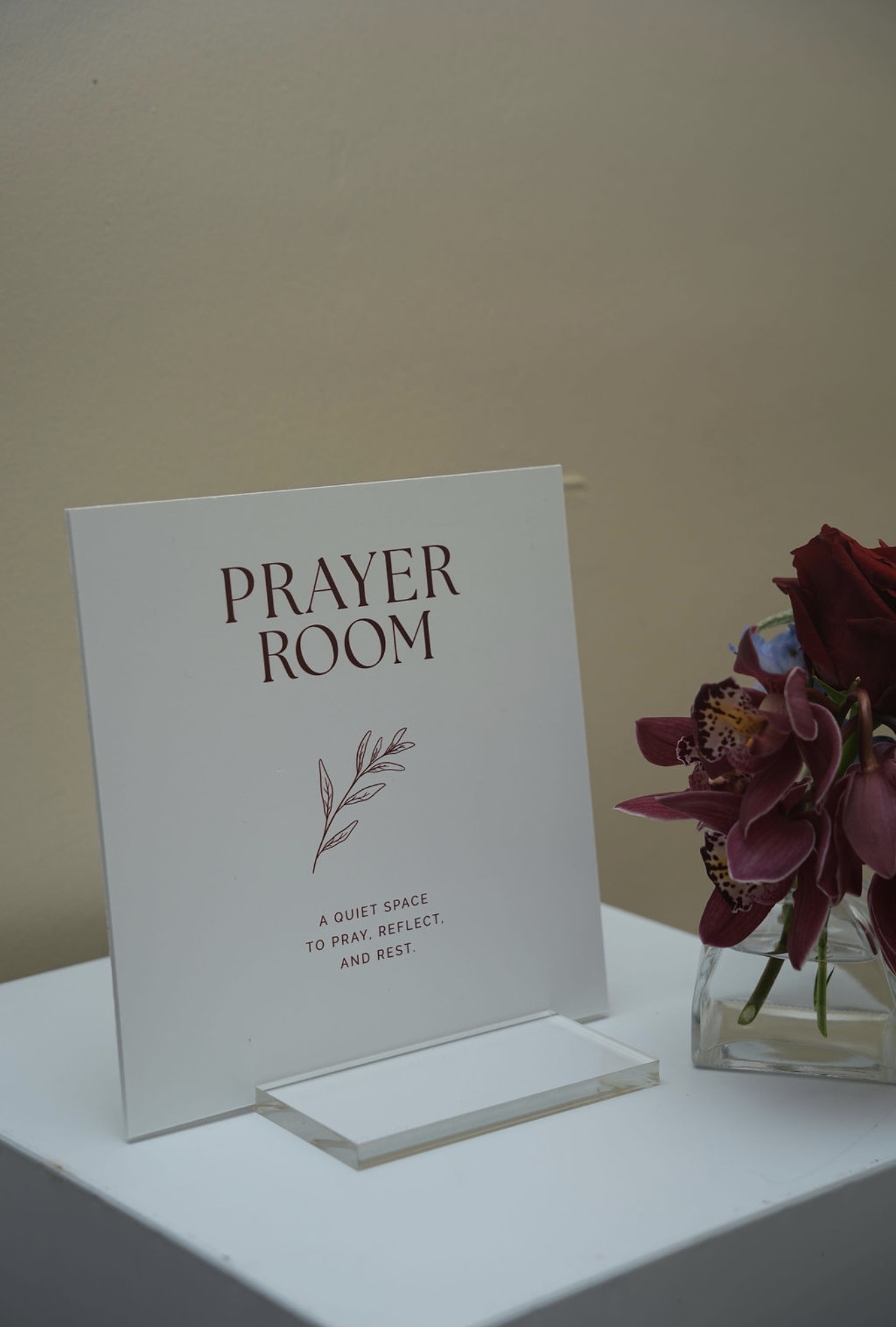 A printed sign reading Prayer Room, a quiet space to pray, reflect, and rest, beside a small vase of burgundy orchids and roses.