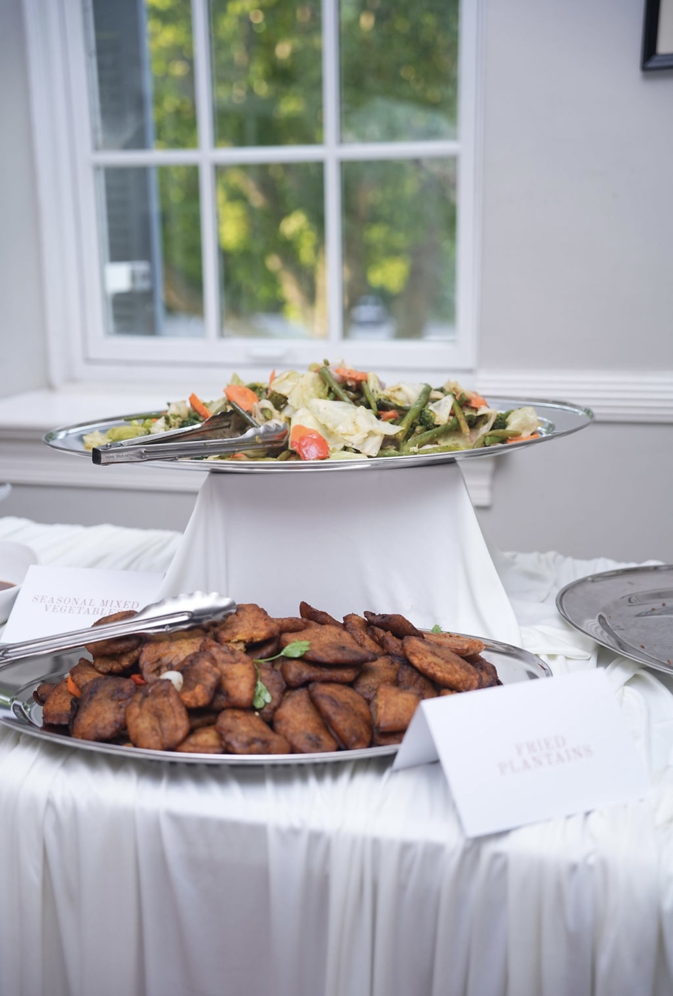 Silver platters of fried plantain and seasonal vegetables on a draped station, each marked with a printed card.