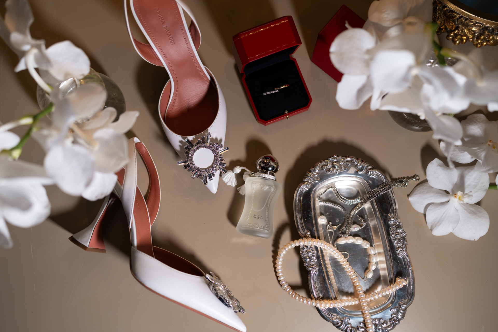 Editorial still life of white bridal shoes, jewellery, a crystal clutch, a ring box and white orchids.
