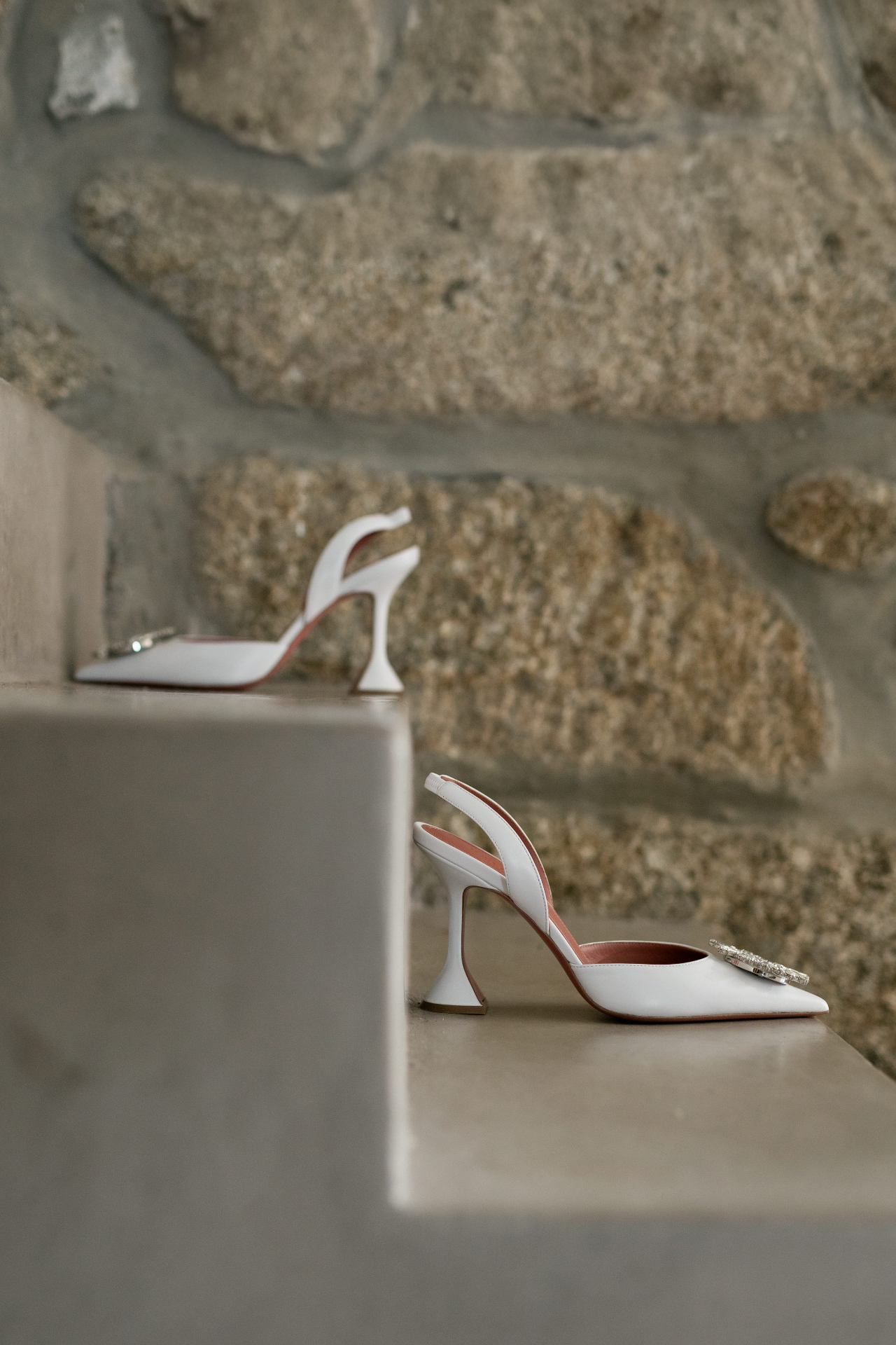 A pair of white bridal heels with crystal detail set on pale stone steps.