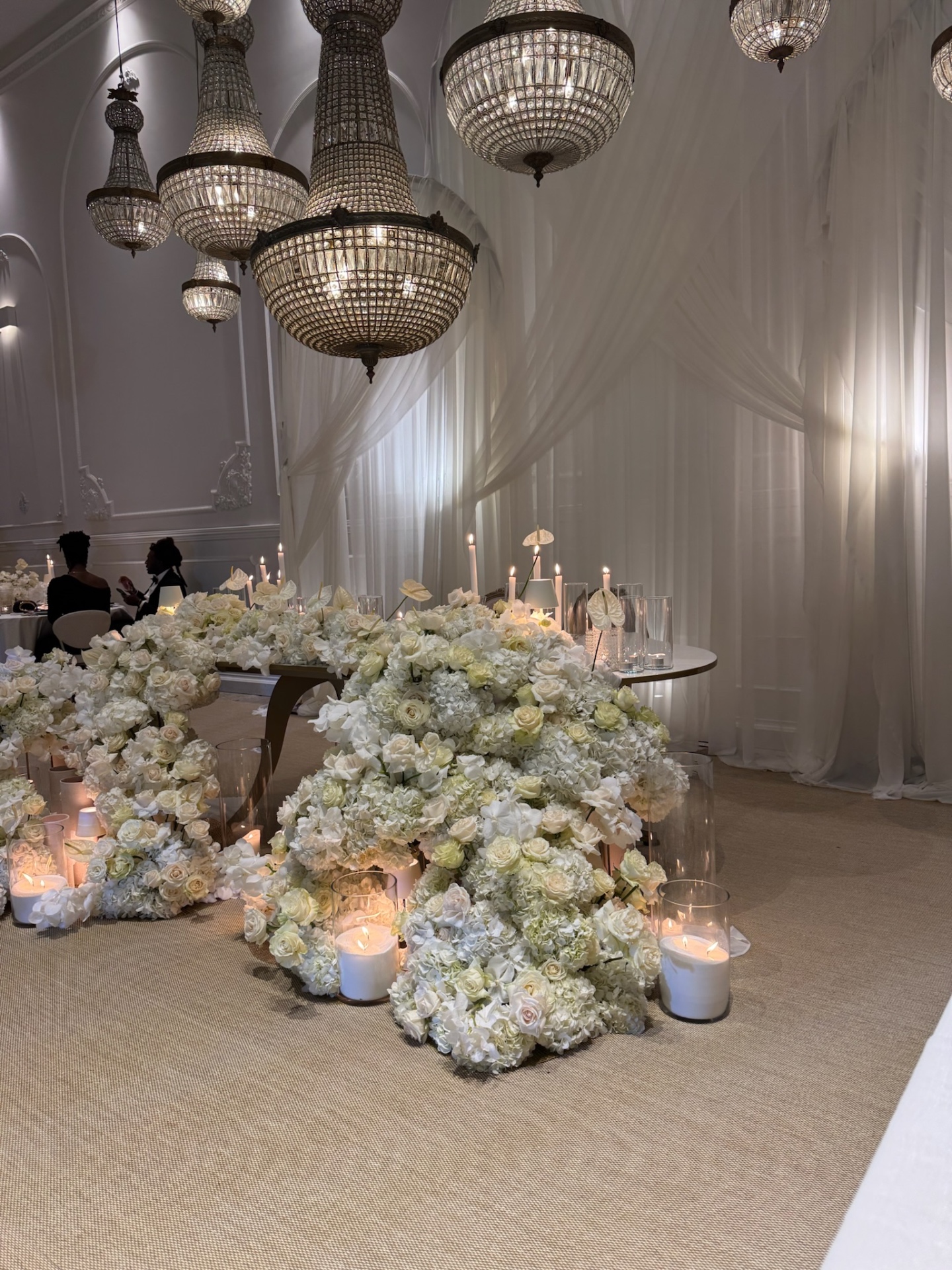 Reception room under crystal chandeliers with a low white floral installation and candlelight.
